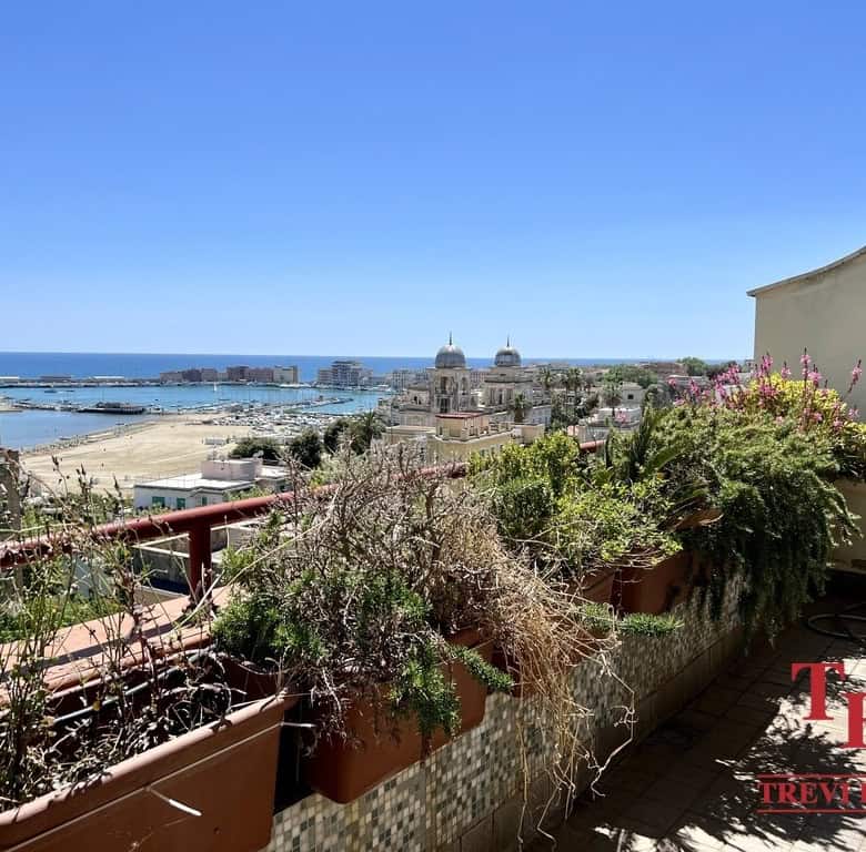 Panoramic view of a luxurious Italian seaside cityscape with historic architecture, lush balcony plants, and the deep blue sea under a clear sky.