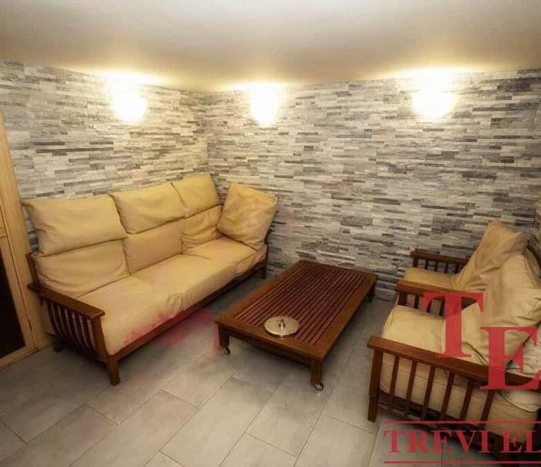 Comfortable beige sofa set with wooden armrests and a matching armchair, situated in a cosy living room with grey stone wall decor and soft lighting, perfect for relaxing or entertaining guests.