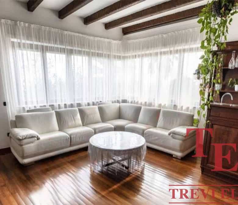Luxurious living room with white sofa and large windows offering plenty of natural light, elegant wooden ceiling beams, and classic interior design by Trevi Elite Italian real estate.