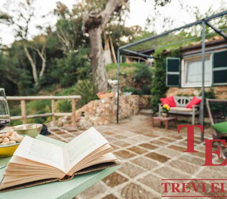 Relaxed outdoor Italian villa terrace with scenic garden view, patio furniture, open book, glass of red wine, and vibrant cushions, perfect for al fresco living.