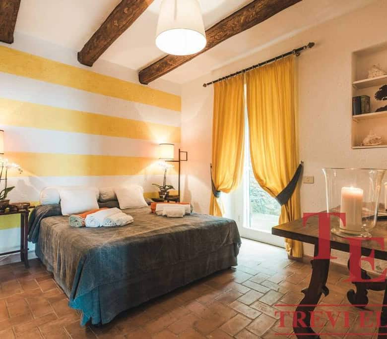 Bright bedroom with warm yellow accents, cozy bed with rolled towels, elegant lighting, and rustic wooden beams, creating a charming Italian home atmosphere.