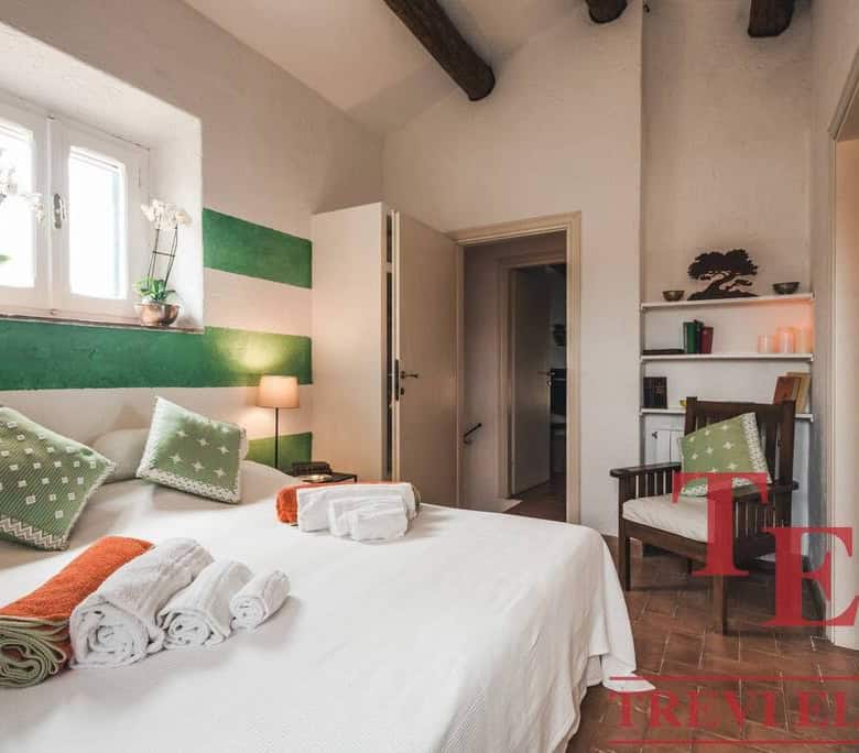 Elegant bedroom with rustic wooden beams and vibrant green accents, featuring a cozy bed with decorative pillows, devotional candles, and stylish furniture for a luxurious Italian residence.