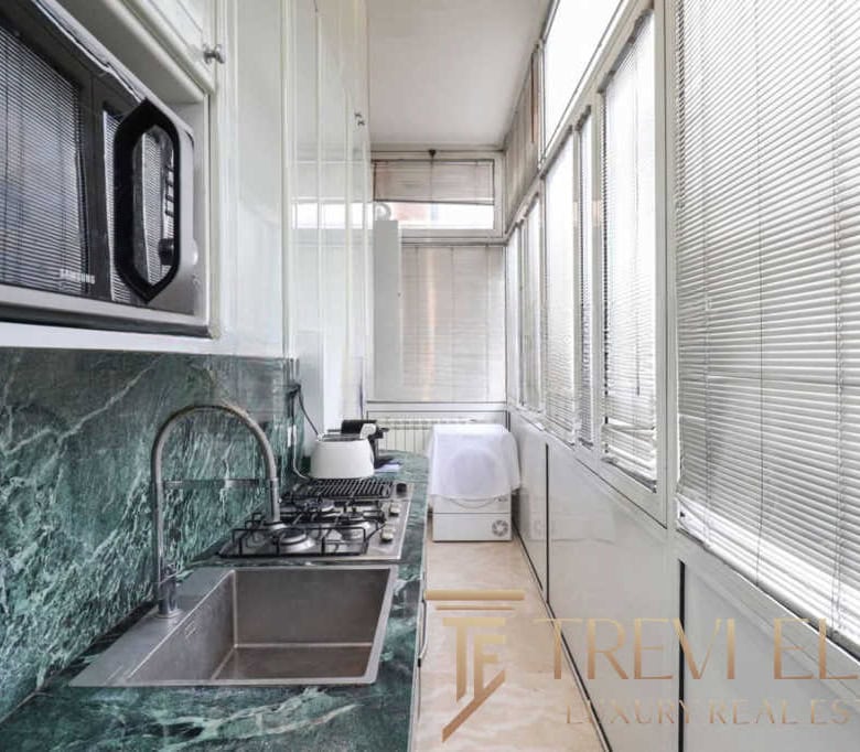 Bright enclosed balcony with green marble countertops, compact stove, microwave, sink, and laundry area, featuring white blinds and ample natural light for functional indoor space.