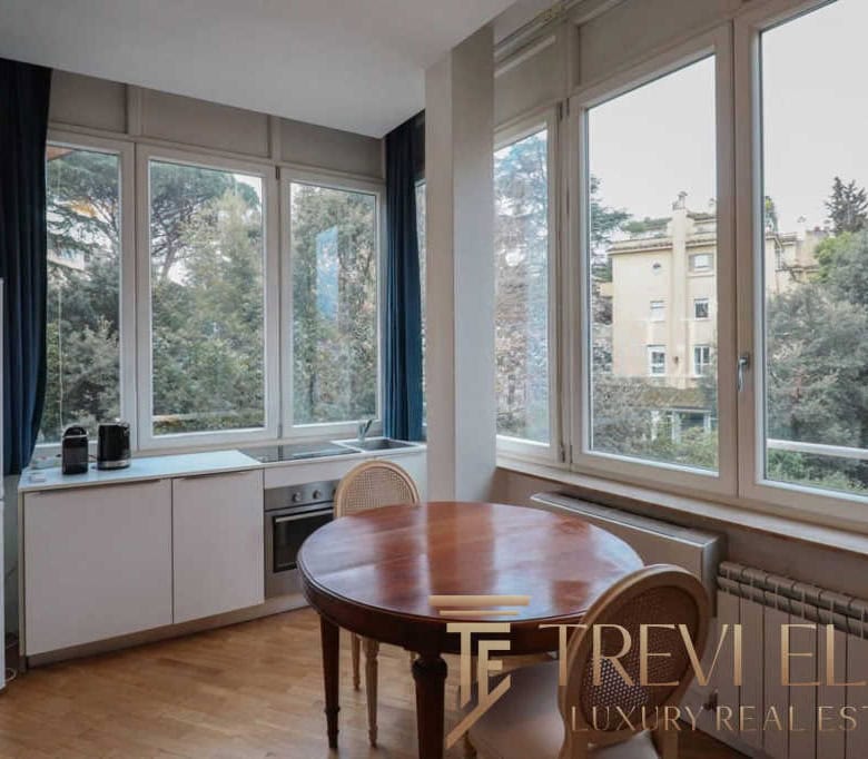 Bright modern kitchen with large windows overlooking greenery, equipped with a refrigerator, oven, and minimalistic white cabinets, creating a spacious and airy environment.
