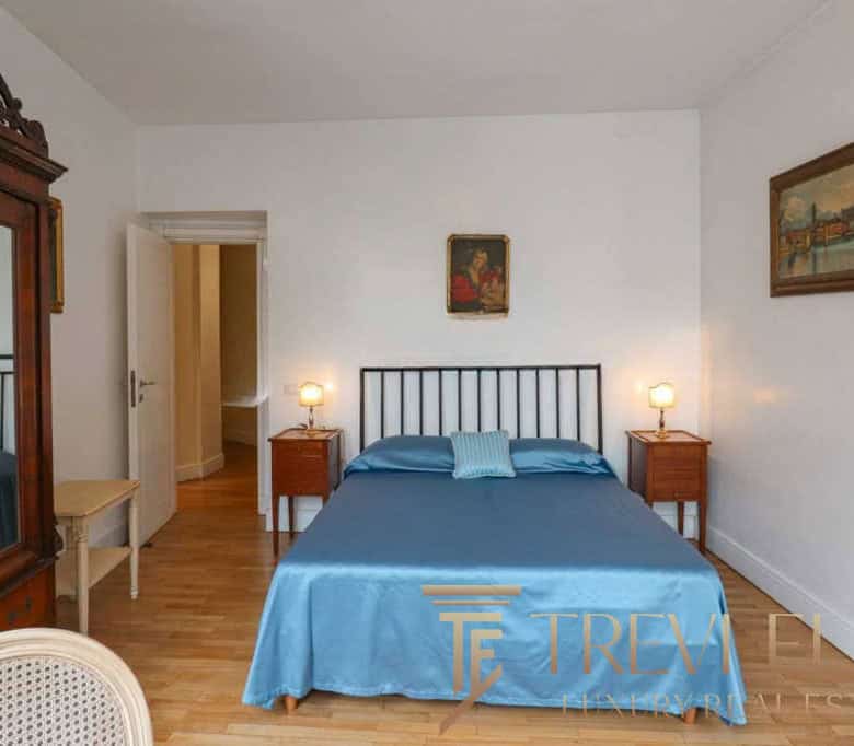 Elegant bedroom featuring a black metal bed frame, blue satin bedding, vintage wooden nightstands with lamps, and classic artwork in a bright, airy space. Perfect for luxurious Italian real estate.