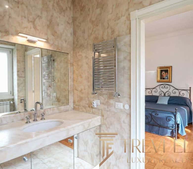 Bright bathroom with large mirror, marble countertop, and modern fixtures leading into a cosy bedroom with wrought iron bed and warm lighting in luxurious Italian estate.