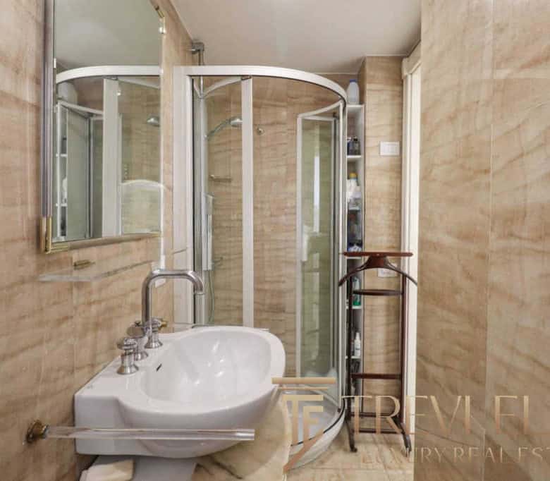 A compact bathroom with a corner shower enclosure, a vintage sink with dual taps, and beige tiled walls in a luxurious Italian apartment.