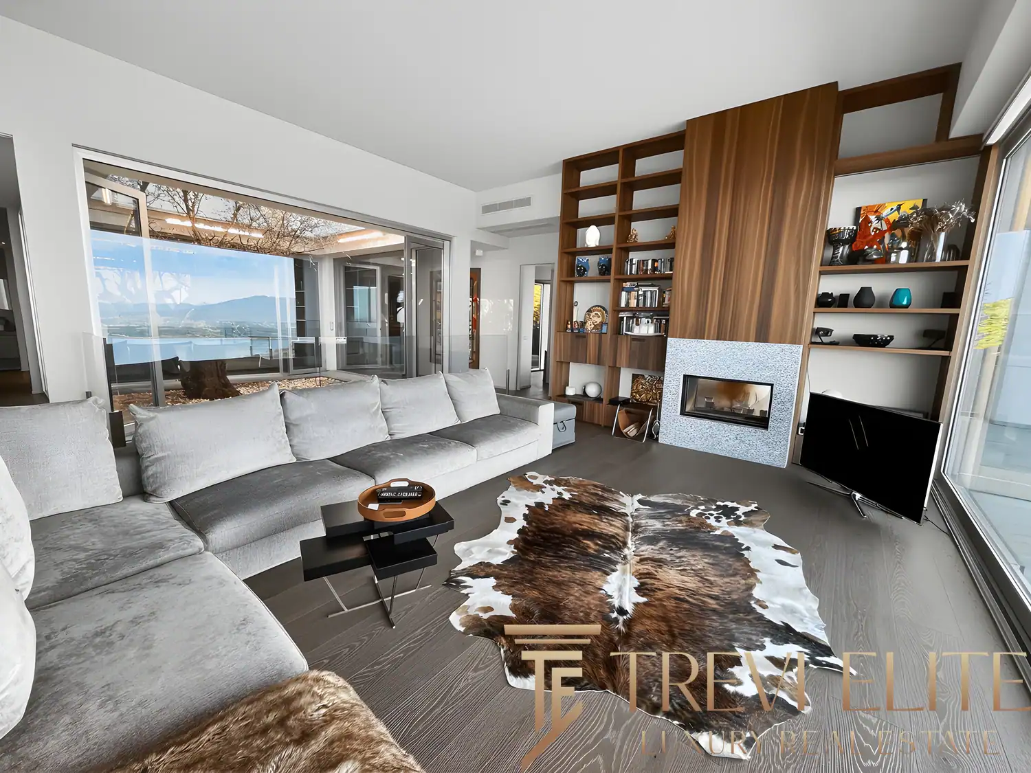 Open-plan living room with floor-to-ceiling glass, grey sofas, and panoramic Lake Maggiore views.